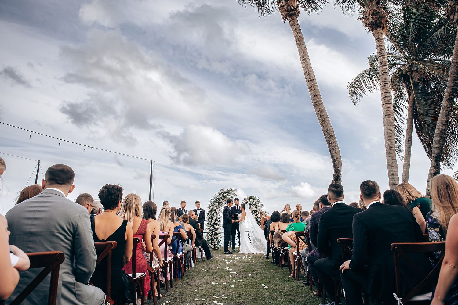 Outdoor wedding ceremony under palm trees with guests seated in rows on a cloudy day.