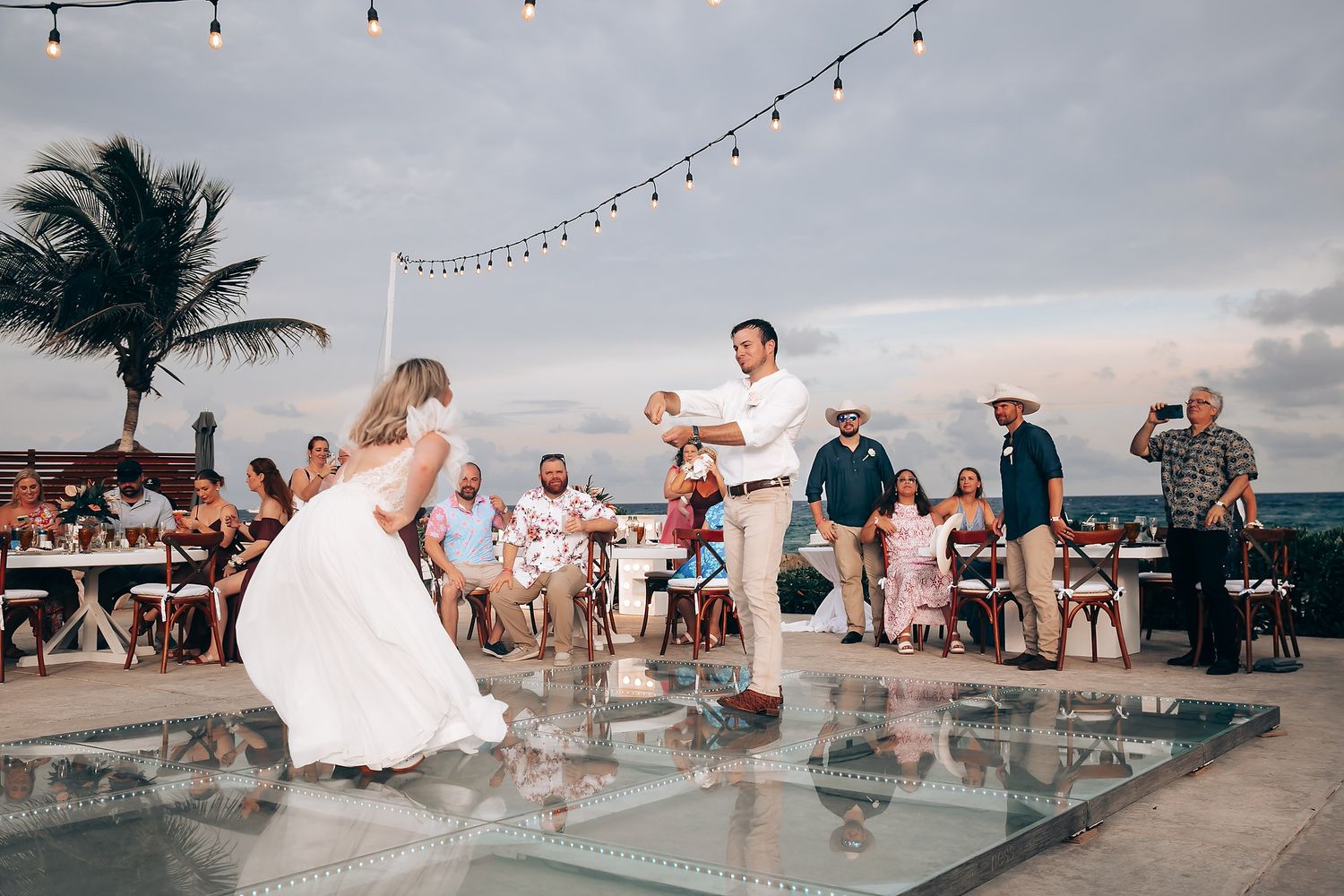 Wedding guests gathered under string lights at an outdoor beachfront reception venue at dusk.