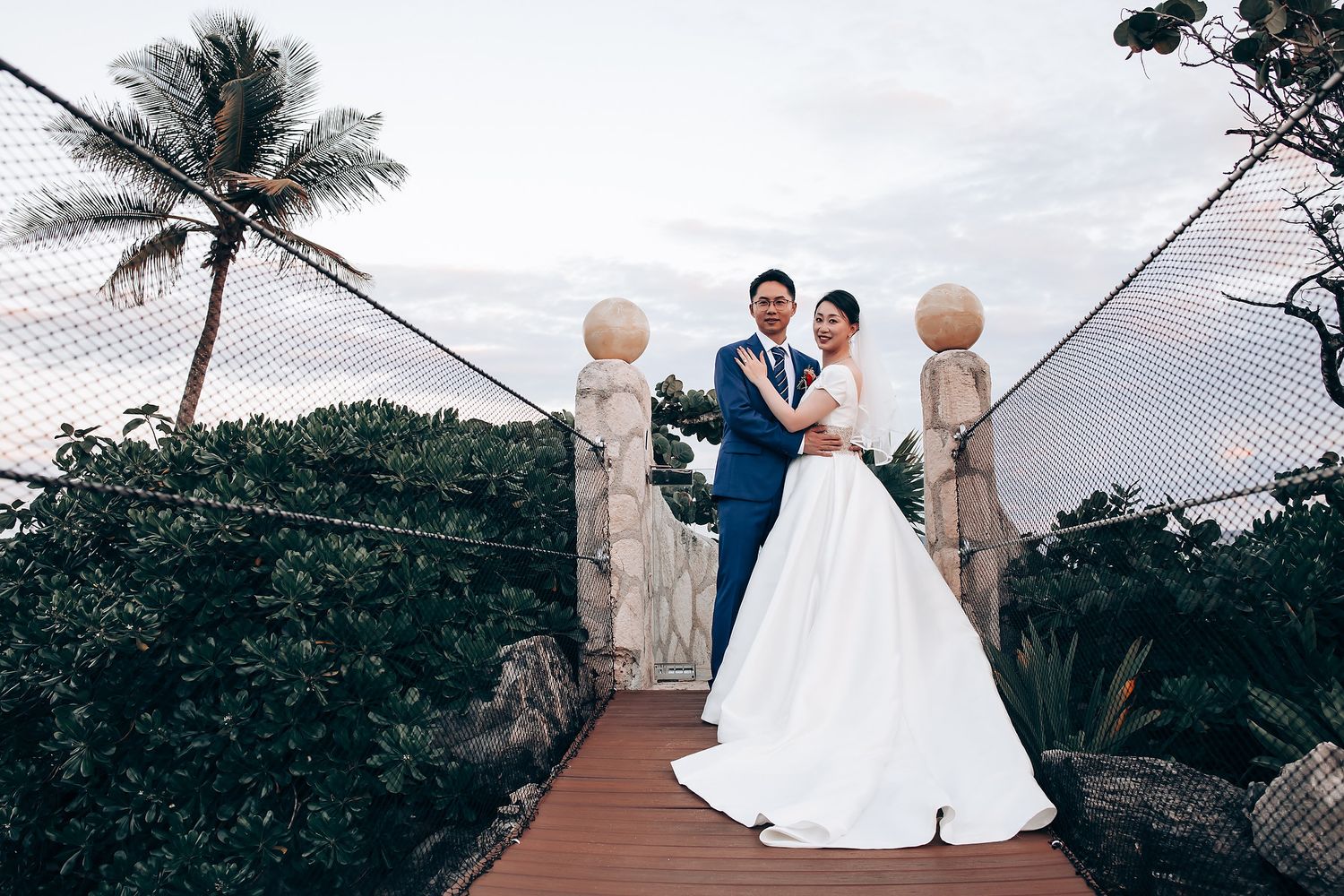 Couple poses on a bridge with palm trees and stone statues at a tropical wedding venue.