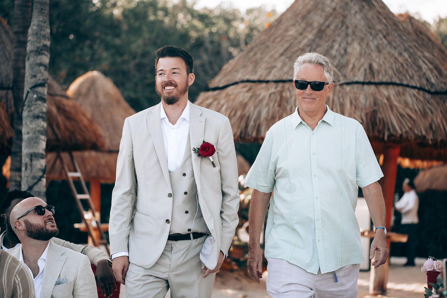 Wedding party in light colored attire walking along a tropical beach with thatched huts.