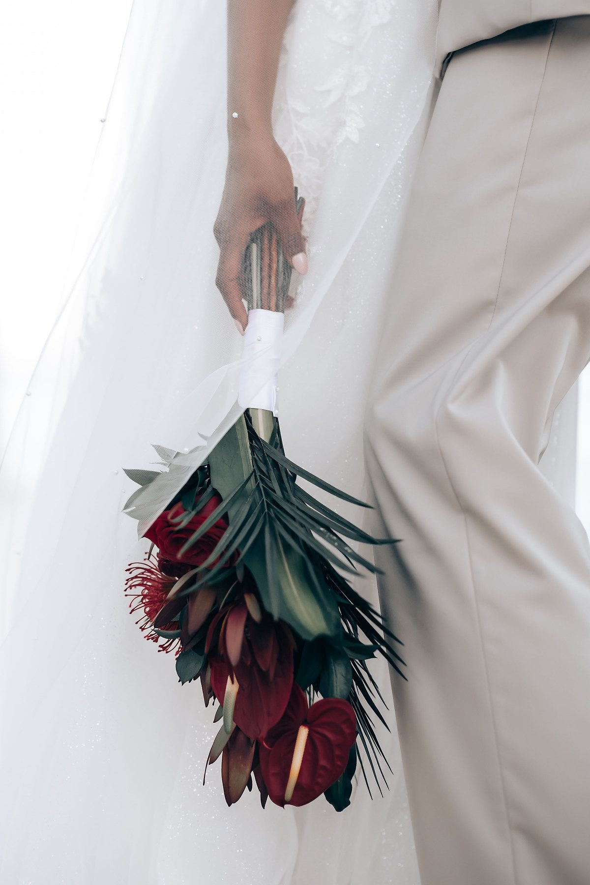 Close-up of deep burgundy and green tropical wedding bouquet against white formal attire.