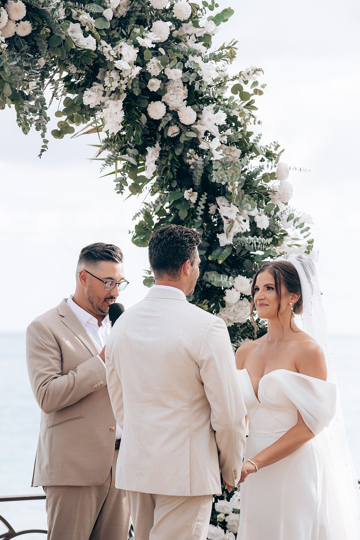 Wedding ceremony under a floral arch with white roses and greenery on a sunny day by the water.