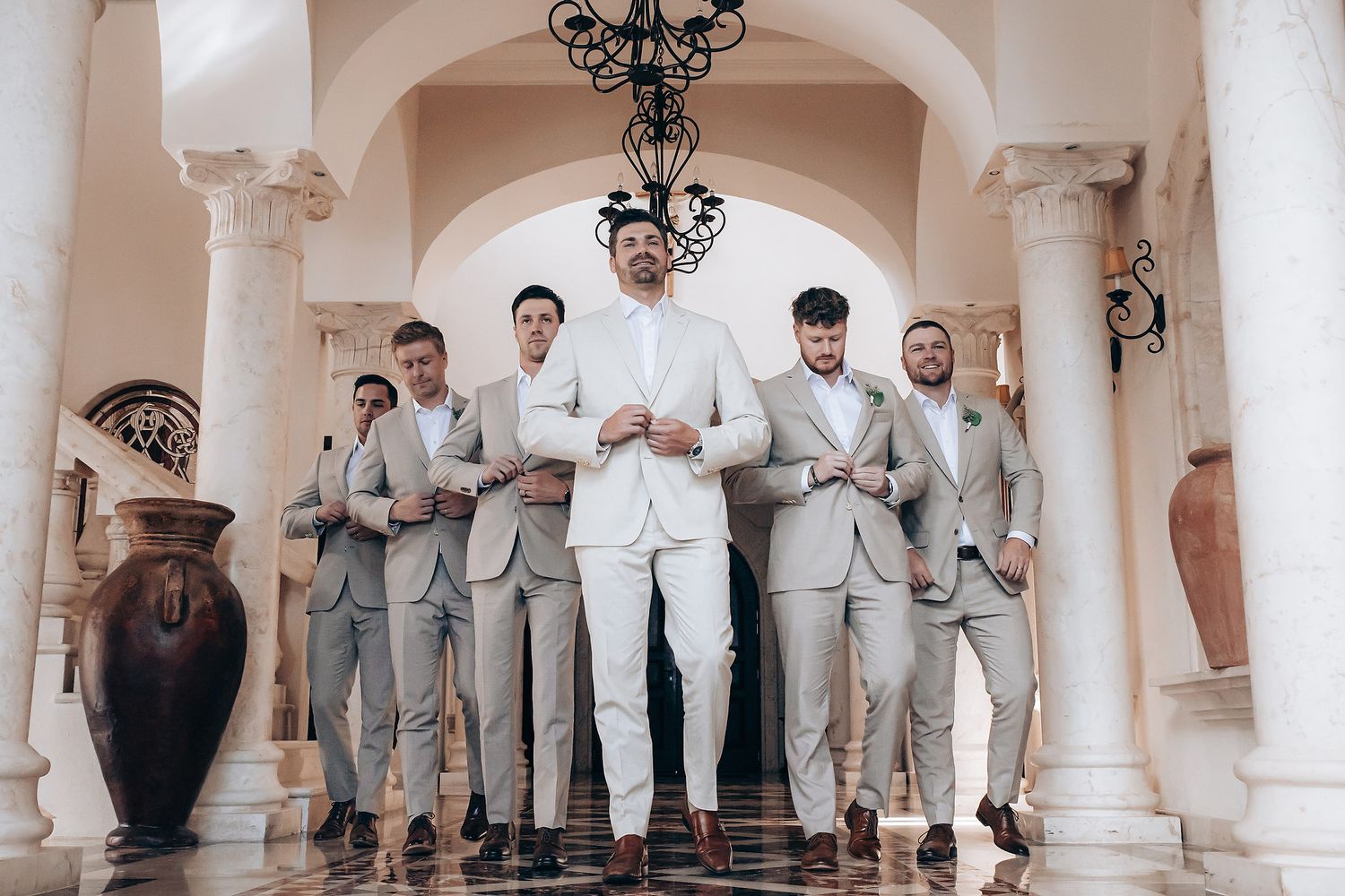 A wedding party wearing light gray and white attire poses together in an elegant arched colonnade.
