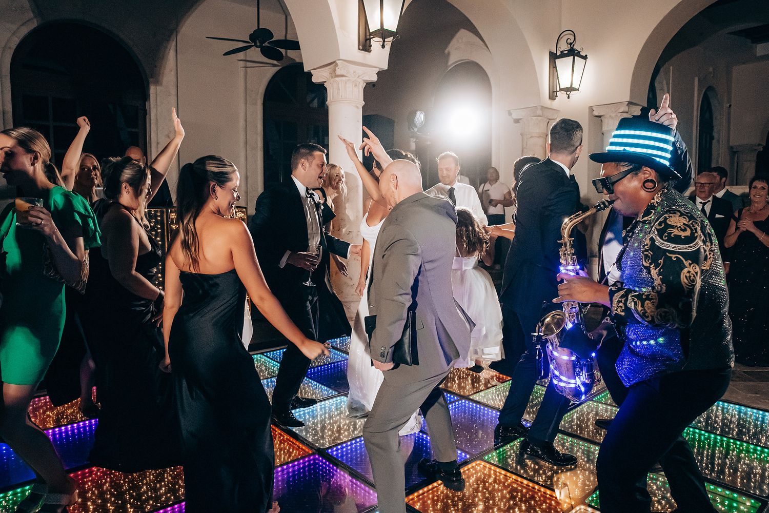 Wedding guests dance and celebrate on a crowded dance floor illuminated by colorful lights and lanterns.