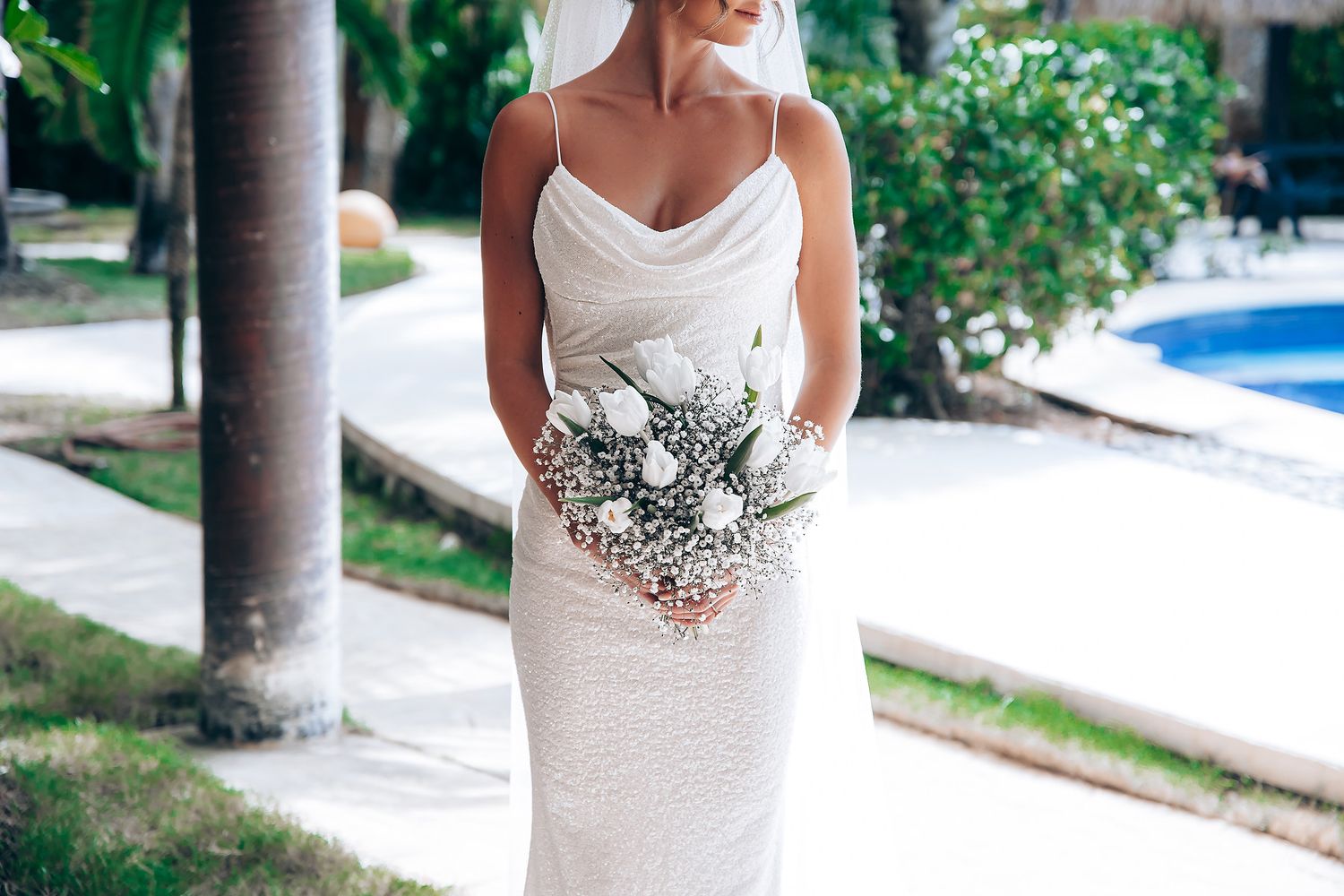 A bride in a white wedding dress holds a bouquet of white flowers while standing between outdoor columns.