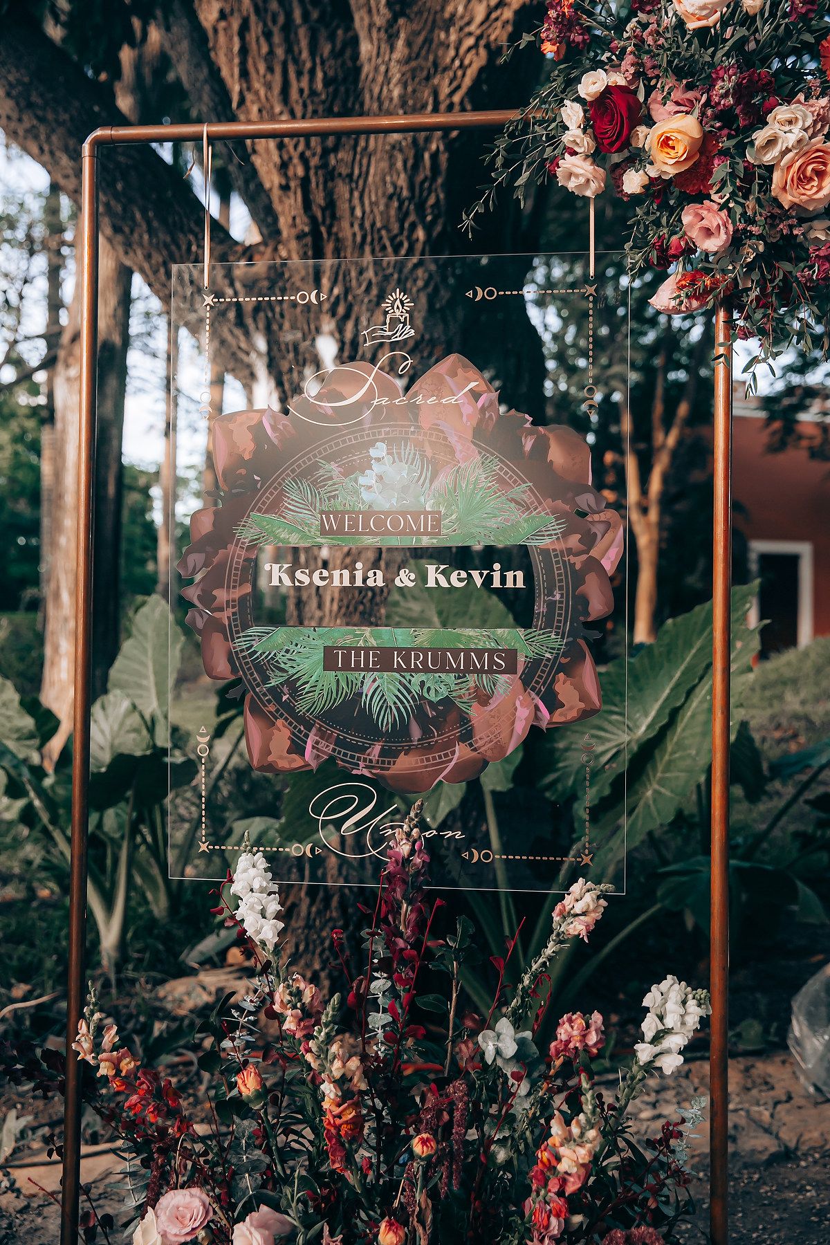 Elegant wedding welcome sign with floral decorations and copper frame in a romantic outdoor garden setting.