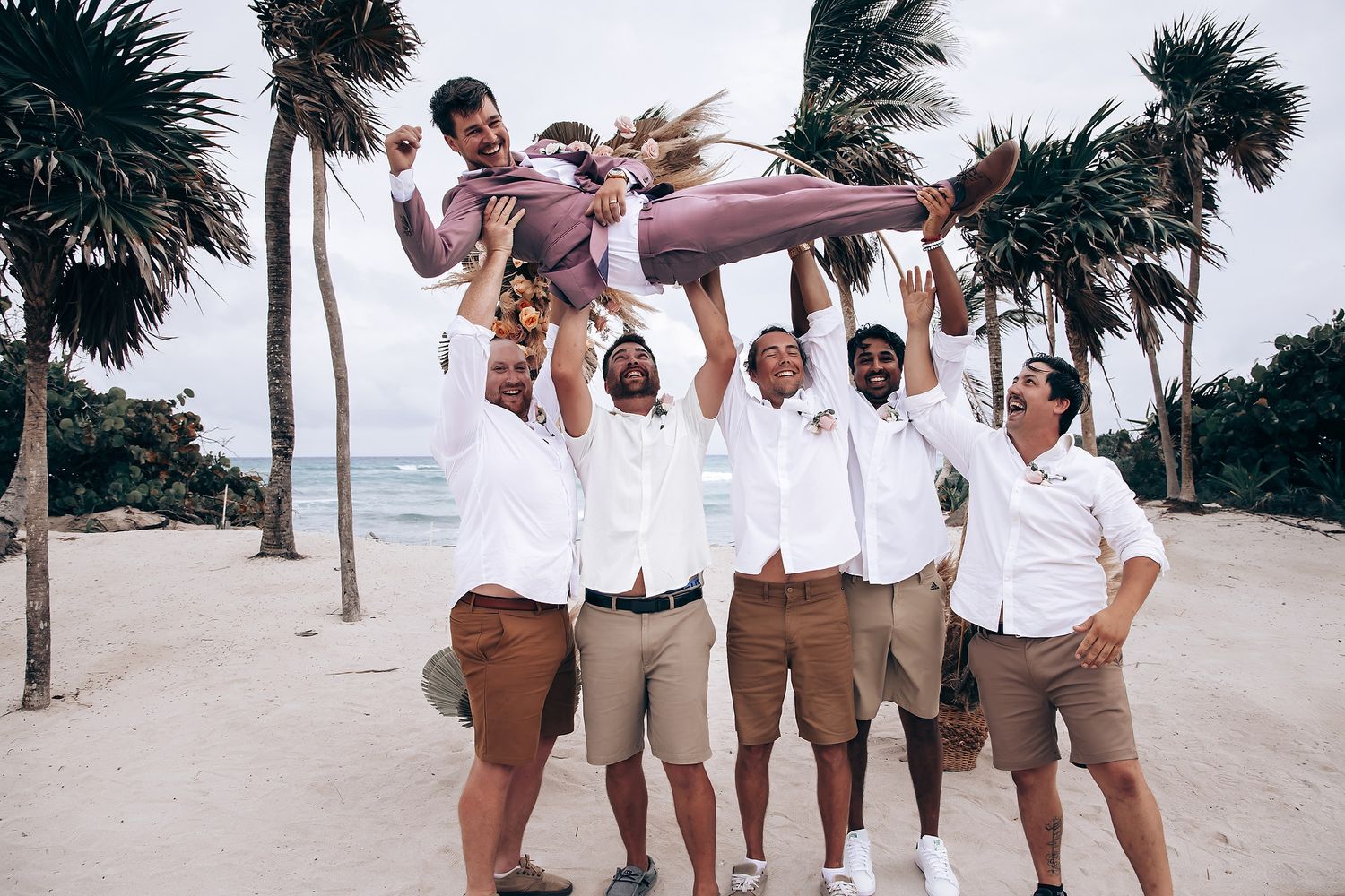 Wedding party members in white shirts and khaki shorts pose playfully on a sunny beach.