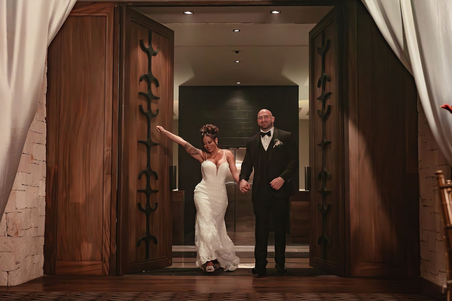 Bride and guests dance energetically in an elegant reception hall with wooden doors and white drapes.