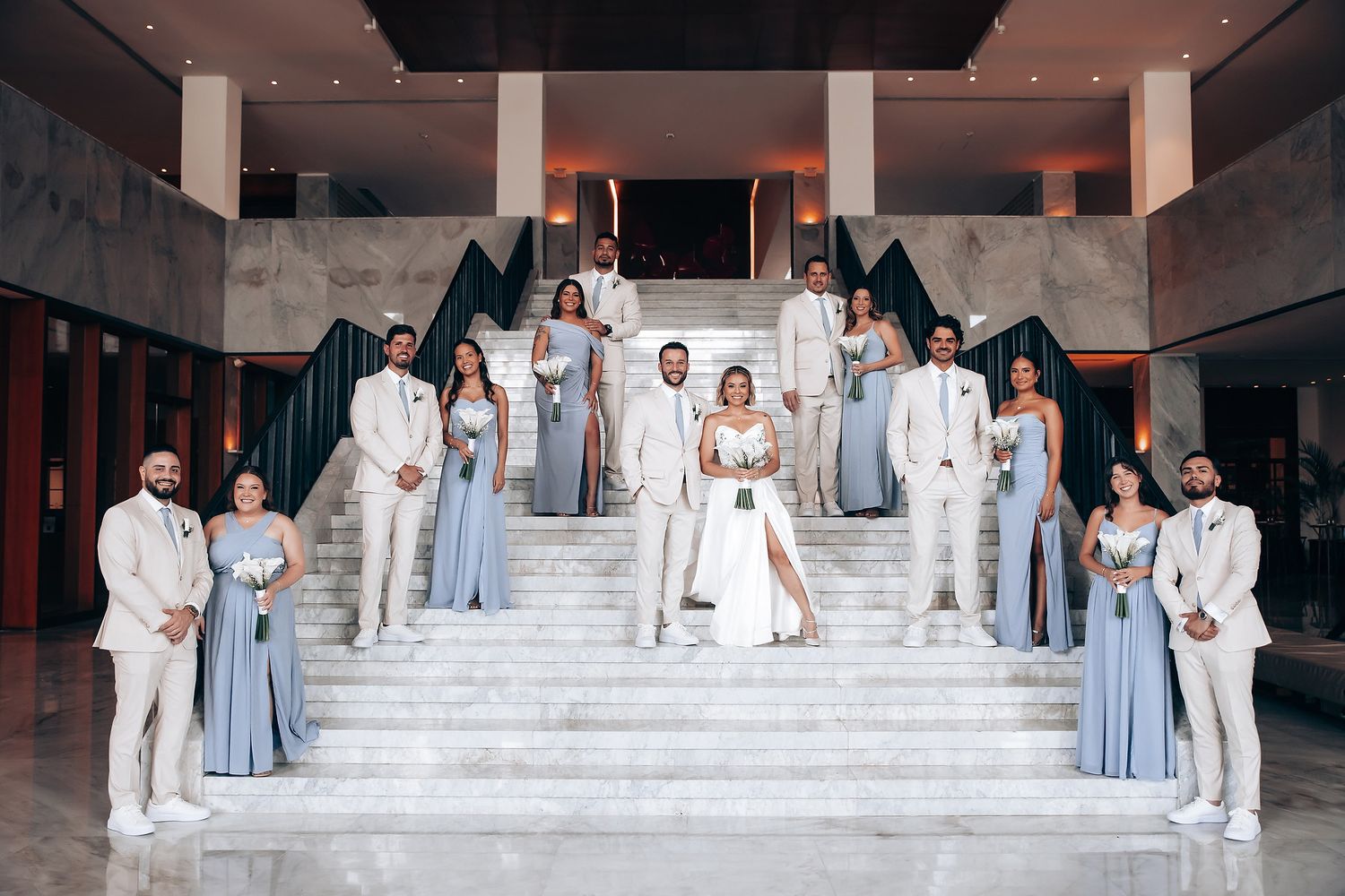 Wedding party dressed in white and light blue poses on grand stone steps for formal group photo.