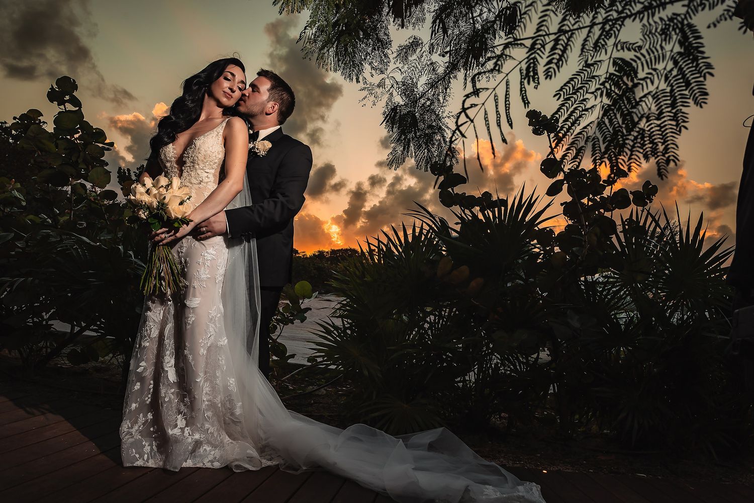 Romantic sunset wedding photos captured against silhouetted palm trees and tropical foliage.
