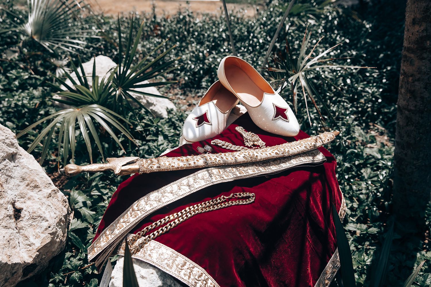Wedding accessories including red velvet shoes and embroidered fabric in outdoor garden setting.