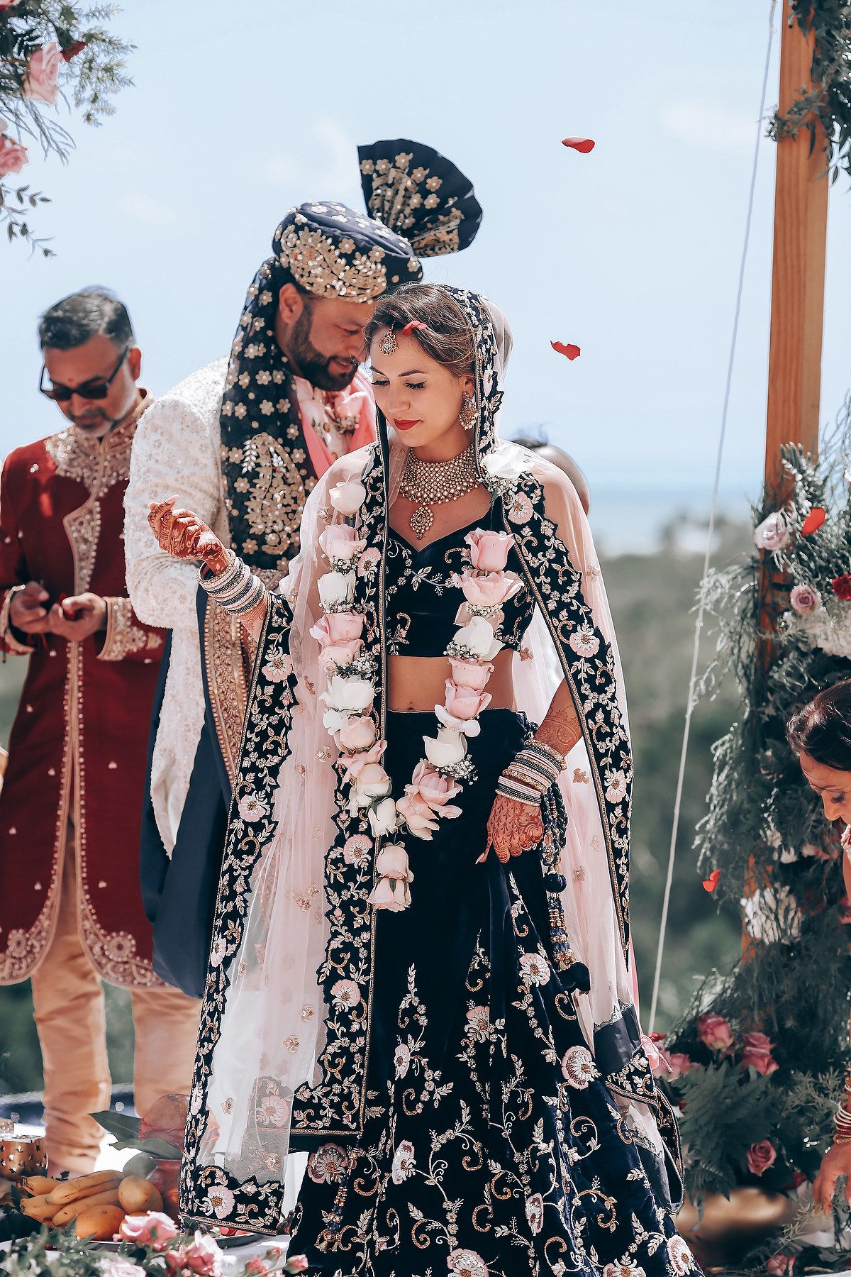 A couple in traditional Indian wedding attire embraces during an outdoor ceremony with floral decorations.