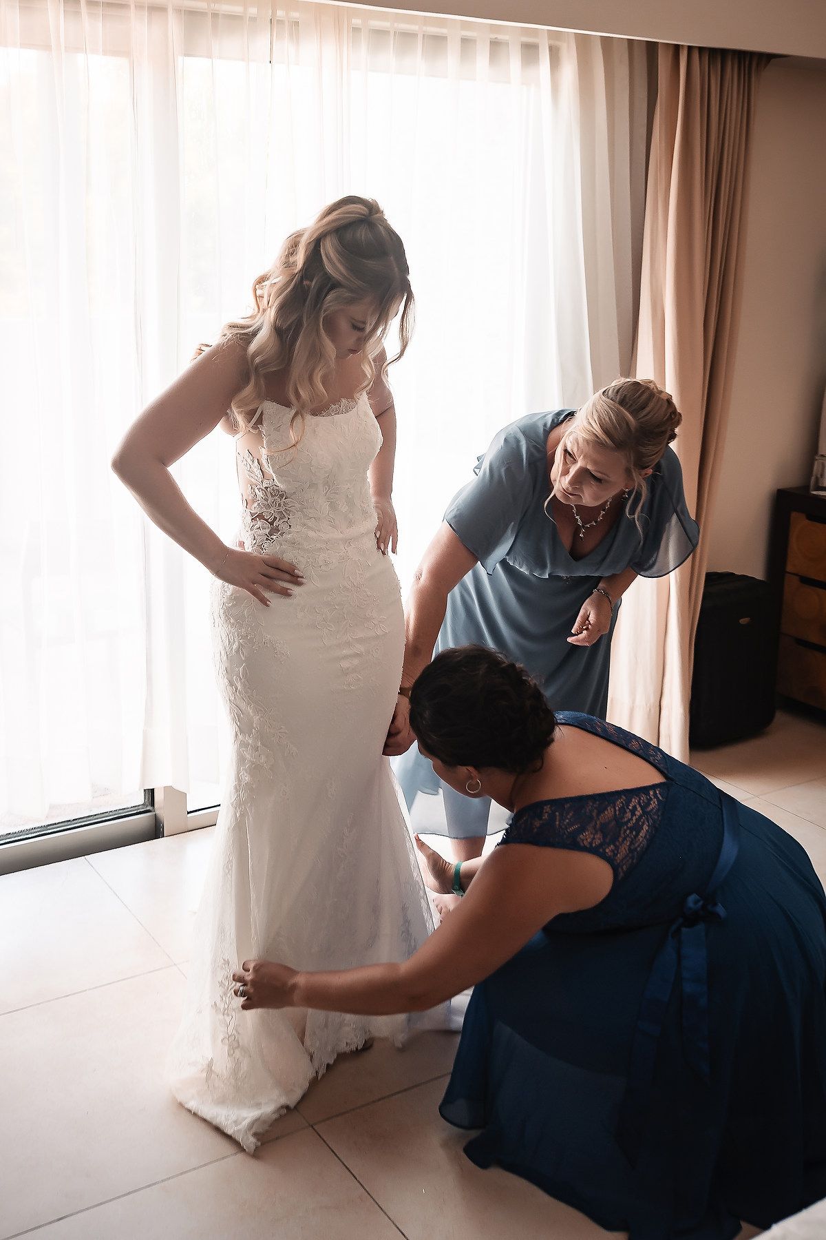 Close-up details of a white lace wedding dress being adjusted in a sunlit room.