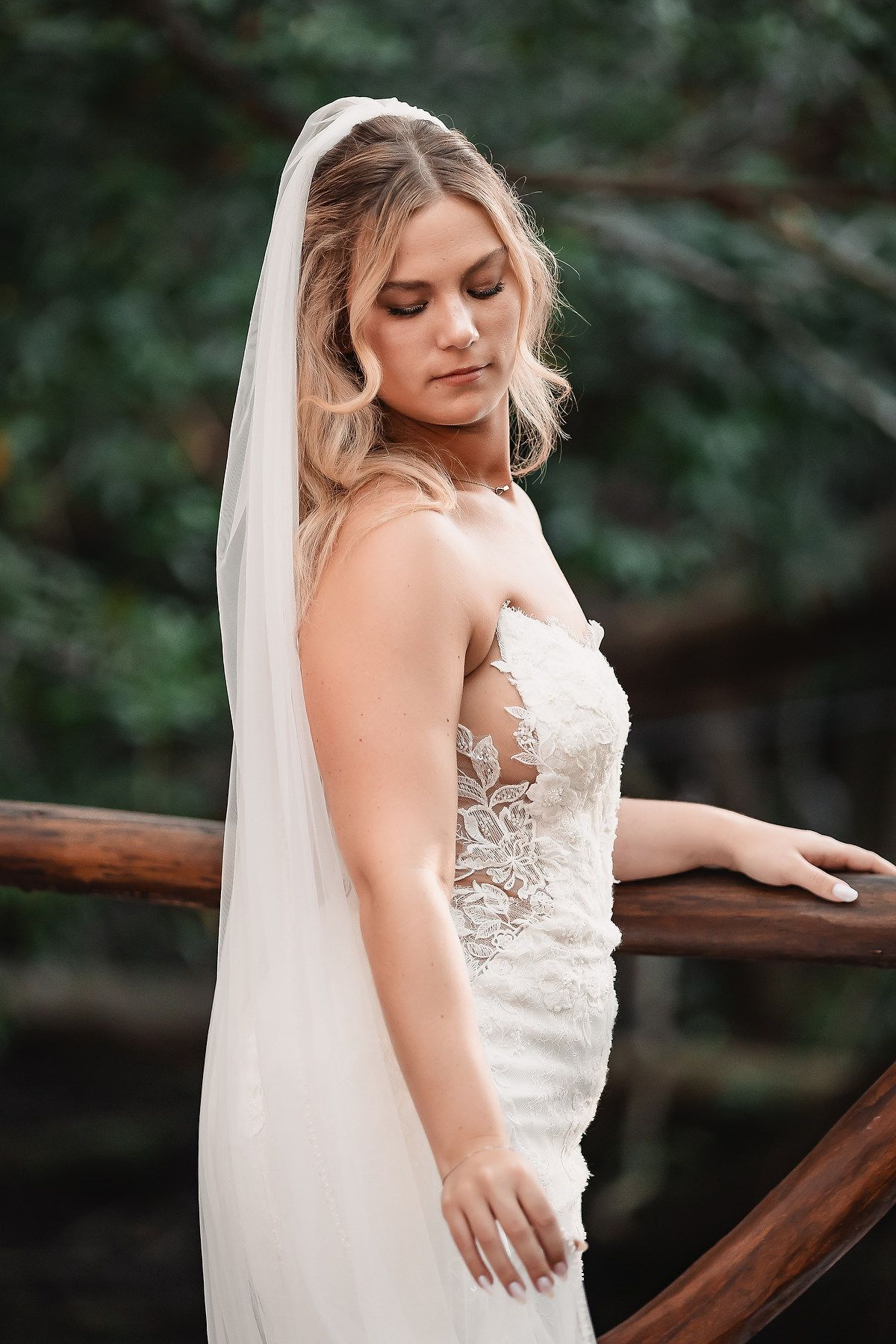 A bride in a lace wedding dress poses elegantly against a wooded backdrop.