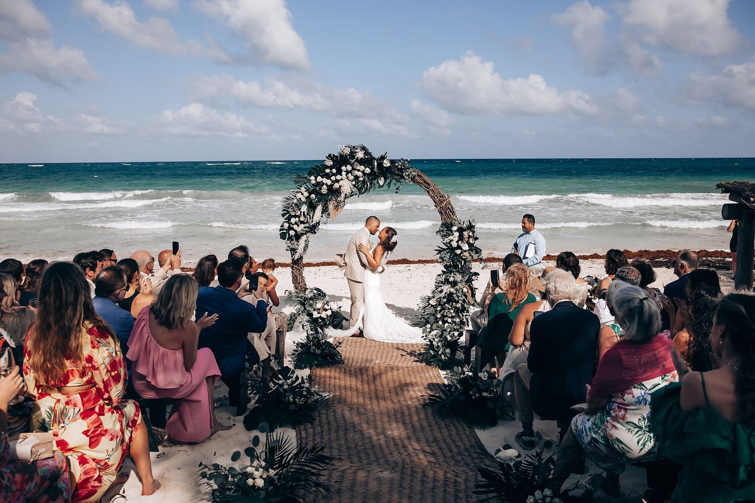 Tropical beach wedding scene with floral arch and seated guests facing the ocean.