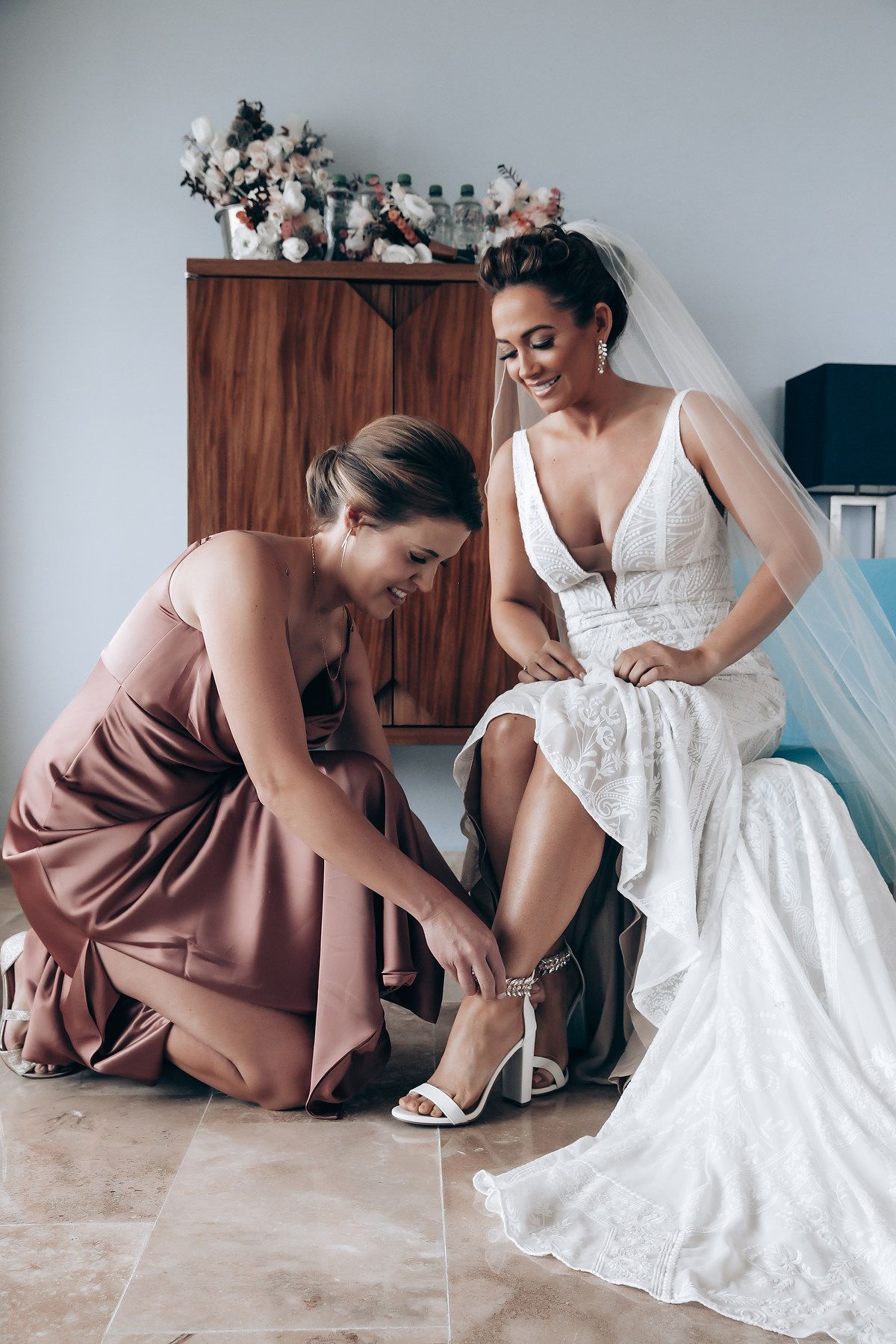 Wedding preparation scene showing a white bridal gown being adjusted with bridesmaids in dusty rose dresses.