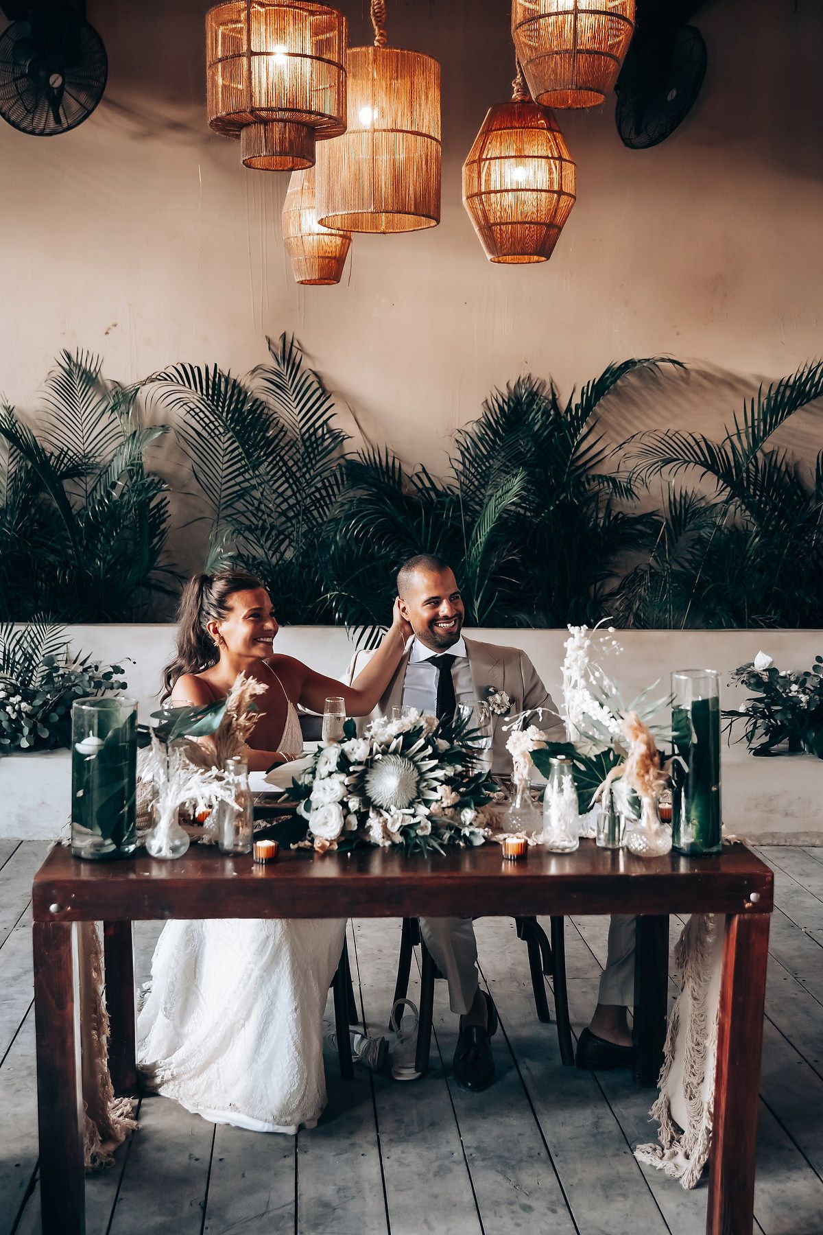 Wedding party seated at a long wooden table with hanging rattan lanterns and tropical palm decor.