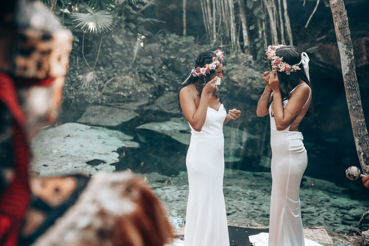 Couple in elegant white dresses standing among trees in an ethereal outdoor setting.