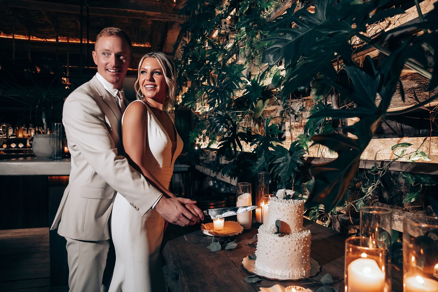 Couple poses with their elegant wedding cake decorated with greenery and candlelight.