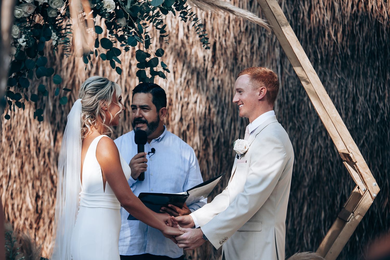 A wedding ceremony takes place under a thatched roof structure with tropical foliage in the background.