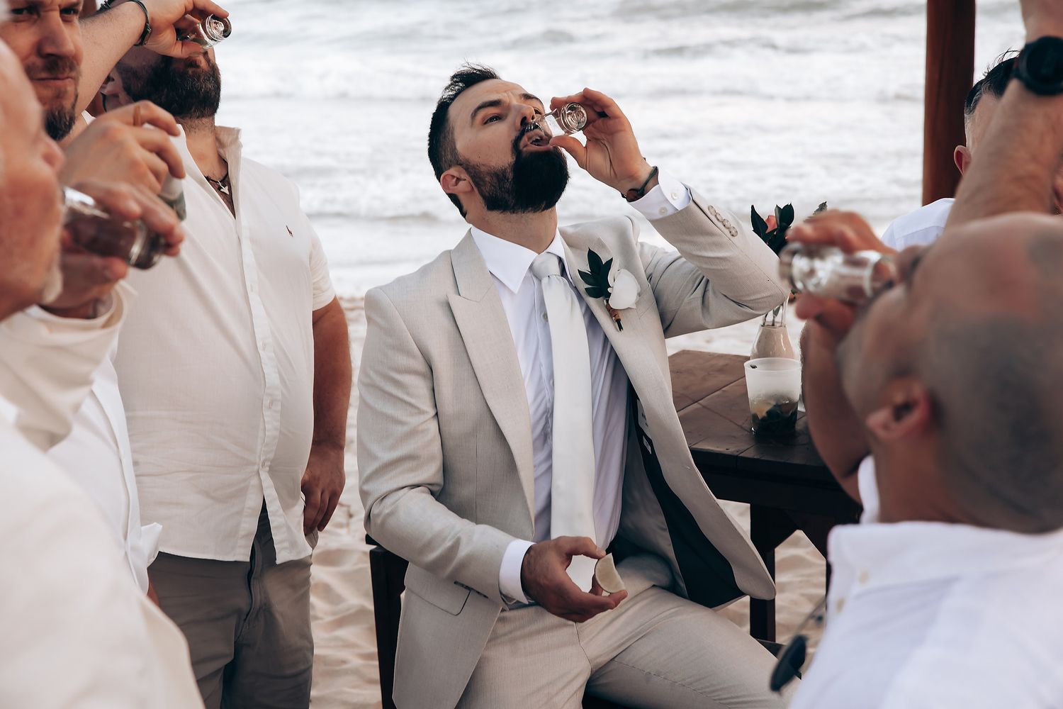 Wedding party celebrating and dancing on beach in light-colored suits.