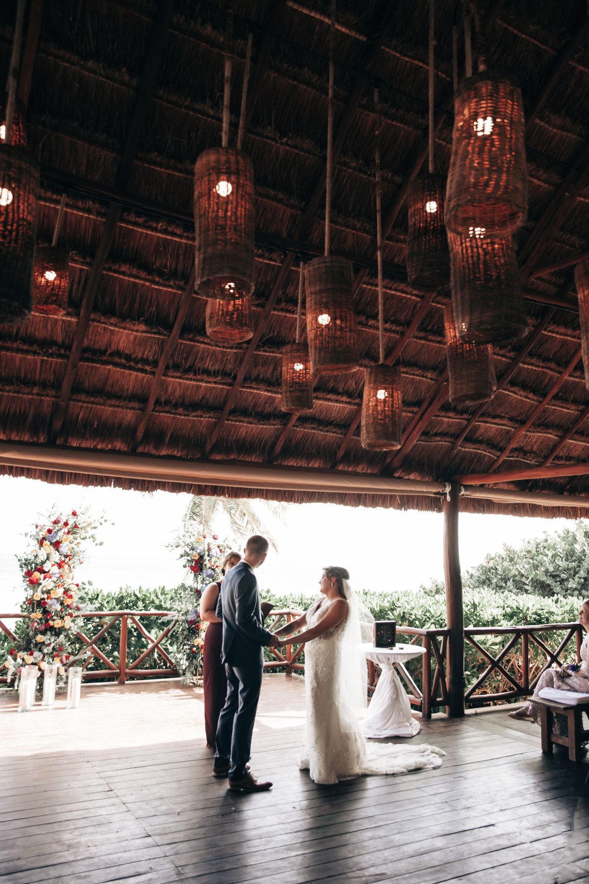 Bride and groom exchanging vows under a rustic thatched roof pavilion with hanging lanterns.