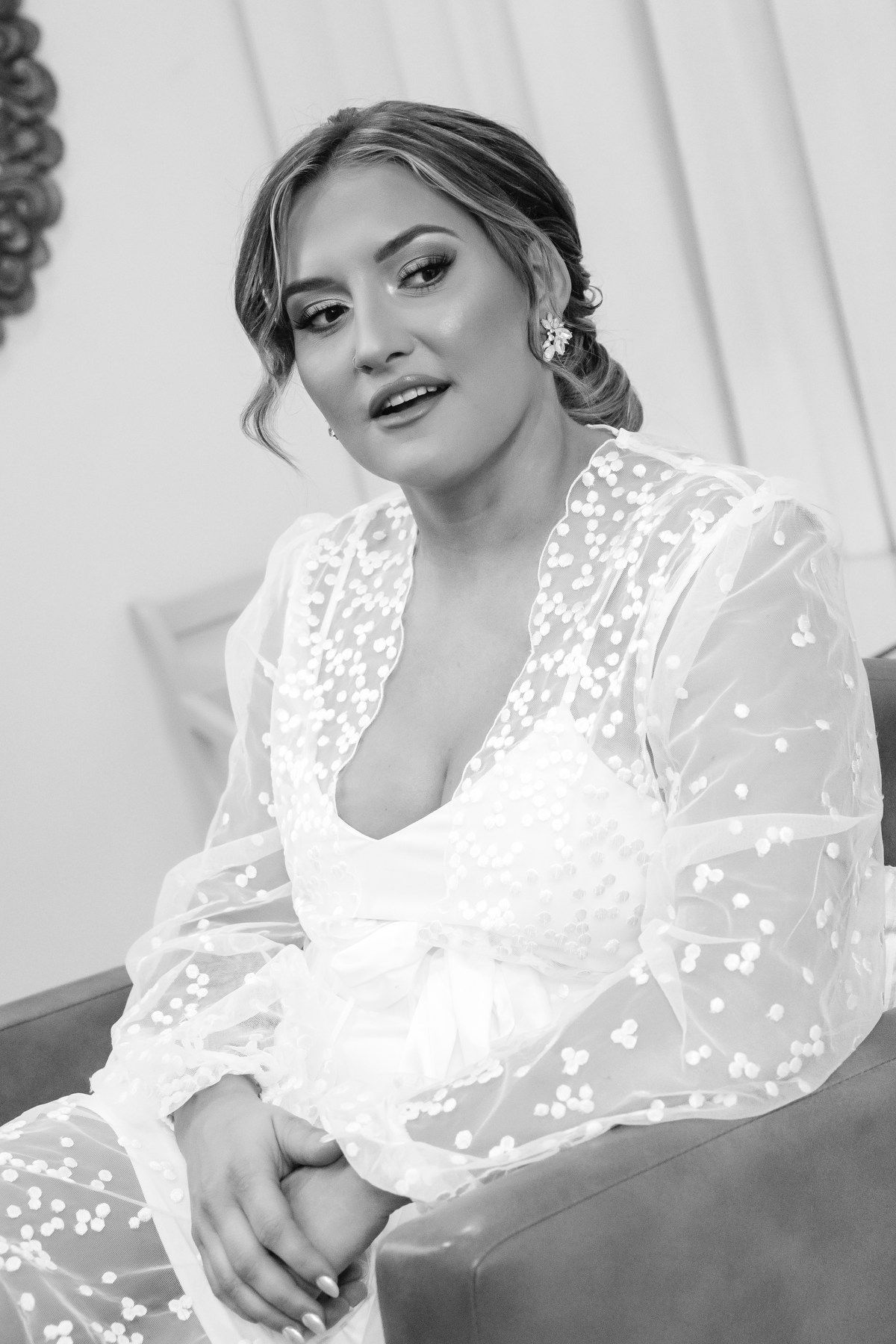 Elegant bride in a sheer floral robe with updo hairstyle and drop earrings sitting gracefully in black and white photo.