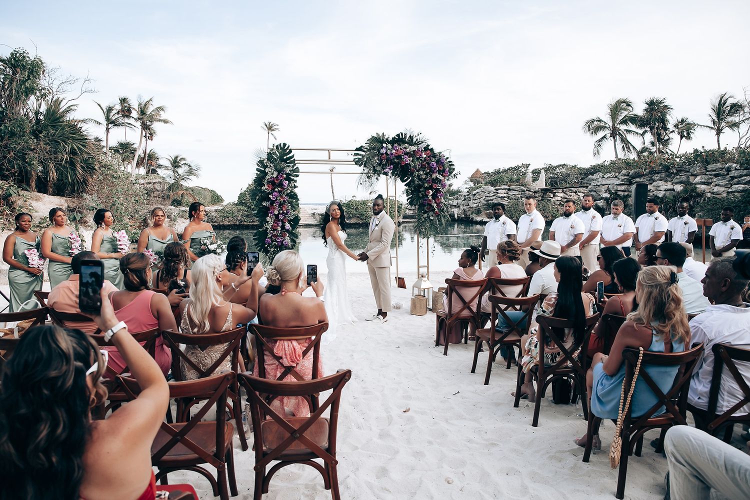 Wedding guests seated in wooden chairs on a tropical beach with palm trees.