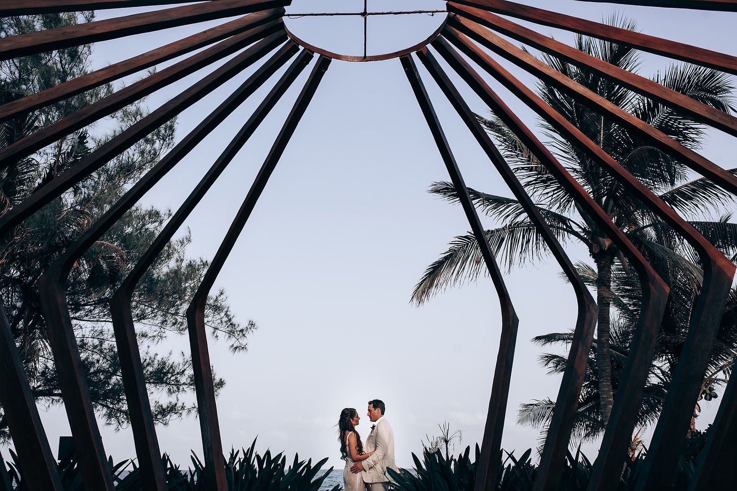 Modern geometric wooden wedding arch with palm trees silhouetted against bright sky.