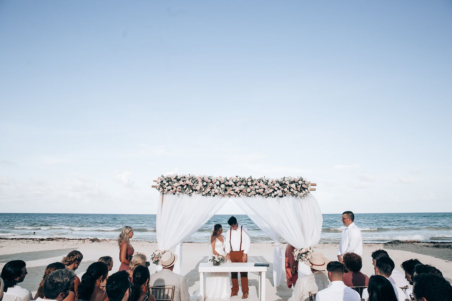 Beach wedding ceremony setup with white chairs and floral arch against blue ocean backdrop.