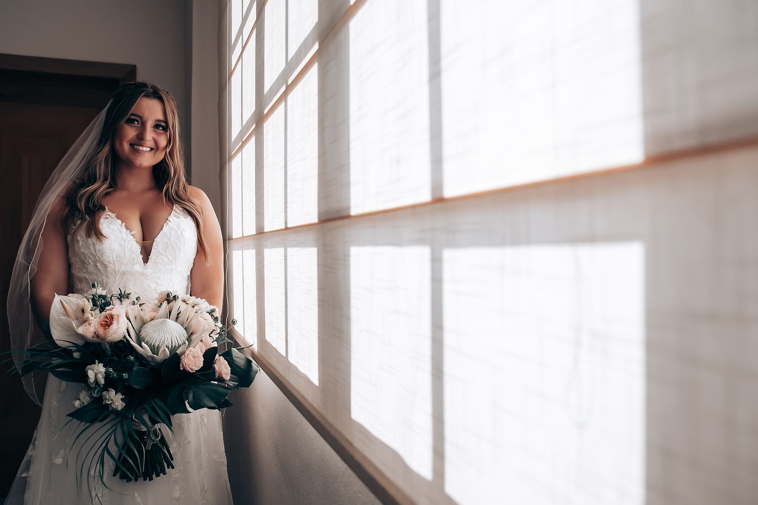 A wedding bouquet of white peonies and roses is held near a sunlit window.