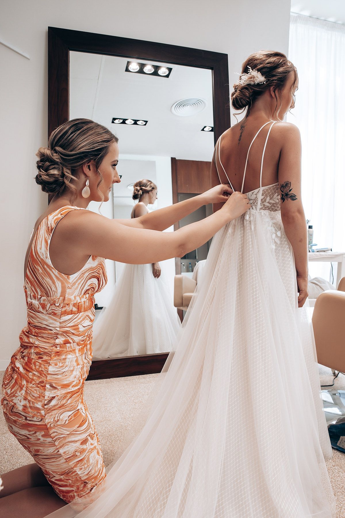 A person in a peach-colored dress helps adjust a flowing white wedding gown train in a bridal preparation room.