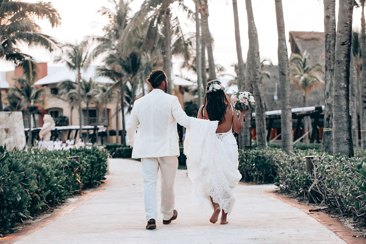 Palm tree-lined pathway with people walking in formal white attire for a beach wedding.