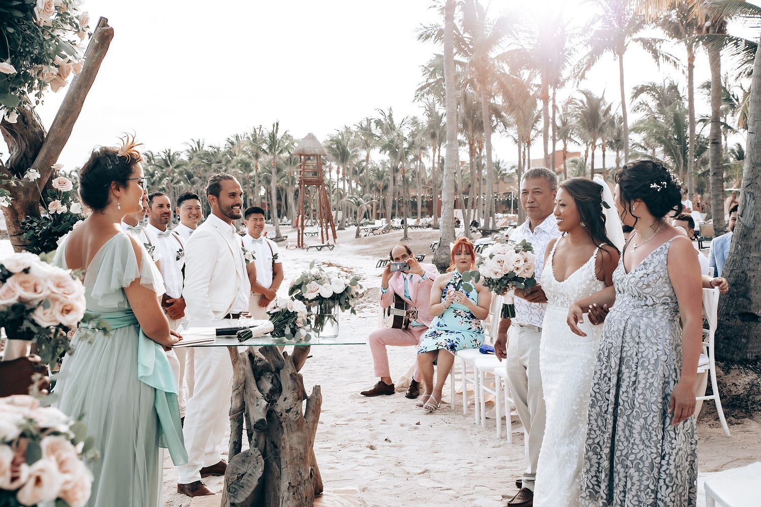 Wedding guests gathered on a sunny beach with palm trees, dressed in light colors for the celebration.