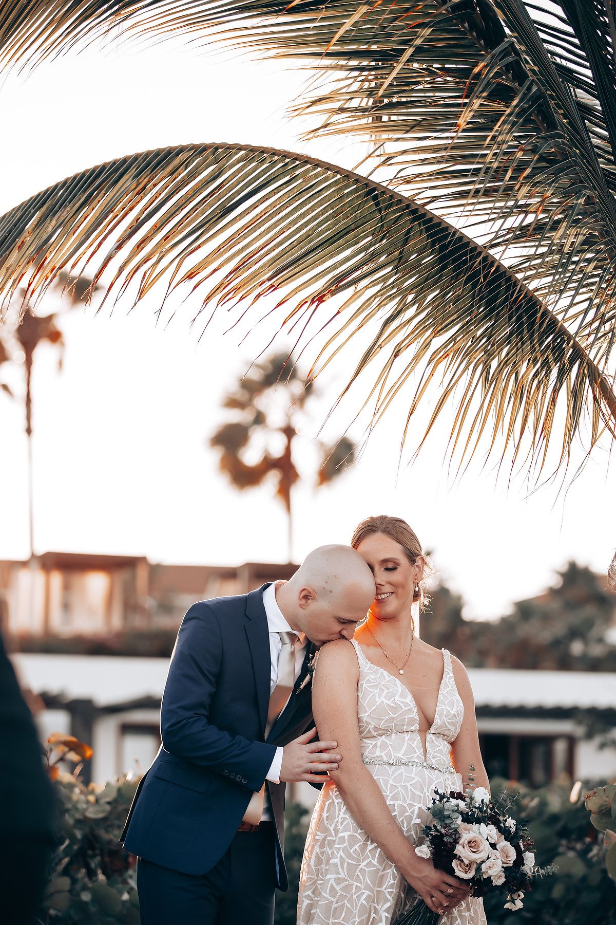 A bride and groom embrace under palm fronds in a romantic outdoor setting.