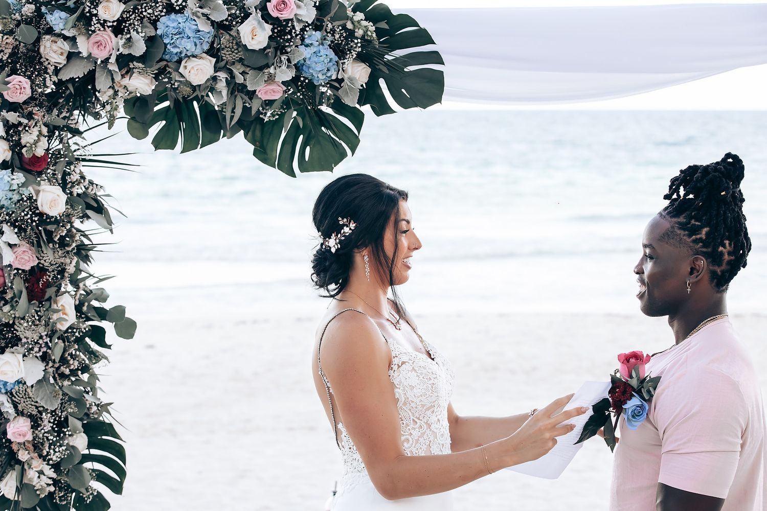 Two women stand together at a wedding ceremony, one wearing a white dress and the other in a pink outfit.