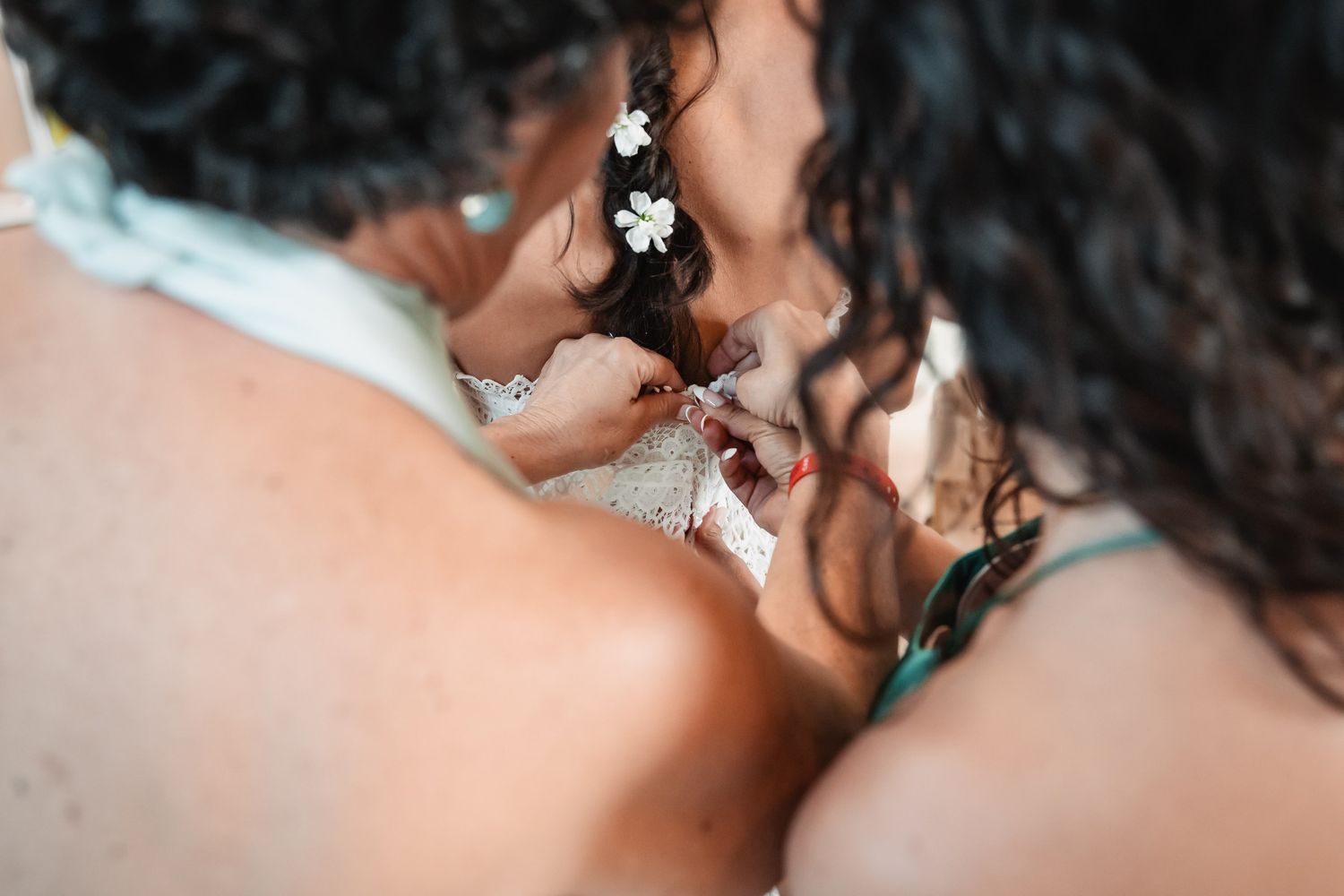 Bridesmaid helping bride with wedding dress details, bride wearing white flowers in dark curly hair.