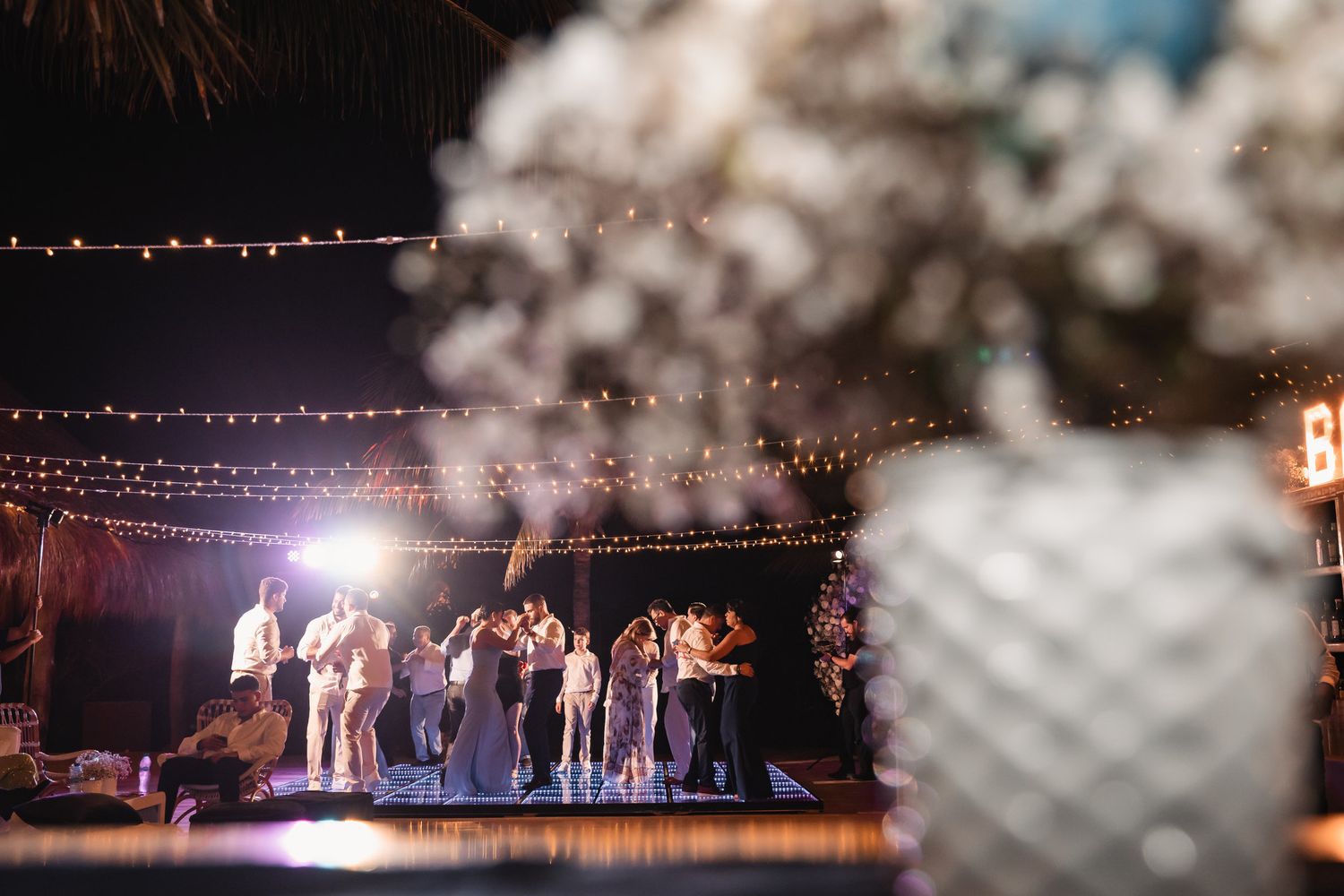 Wedding guests dancing on illuminated dance floor with string lights and white floral centerpiece in foreground.