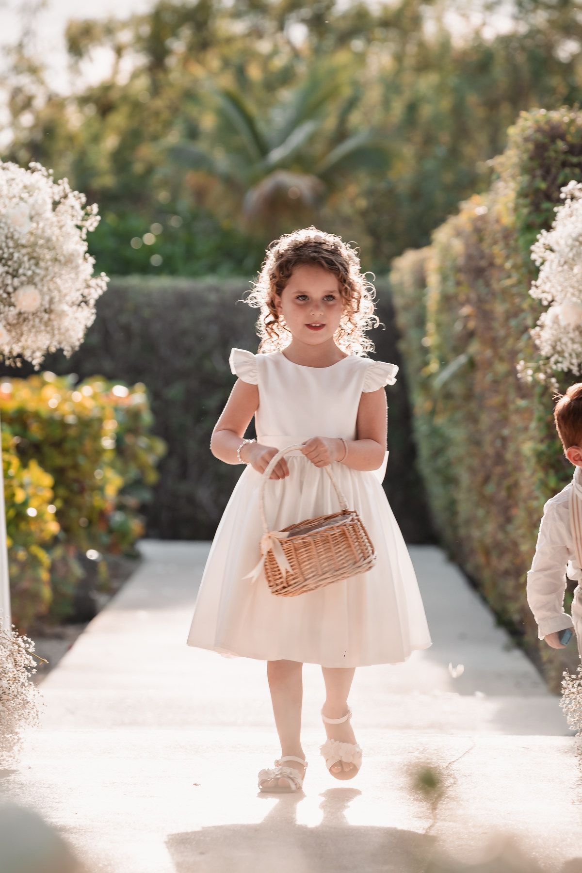 A flower girl in a white dress carries a wicker basket down a sunlit garden aisle at a wedding.