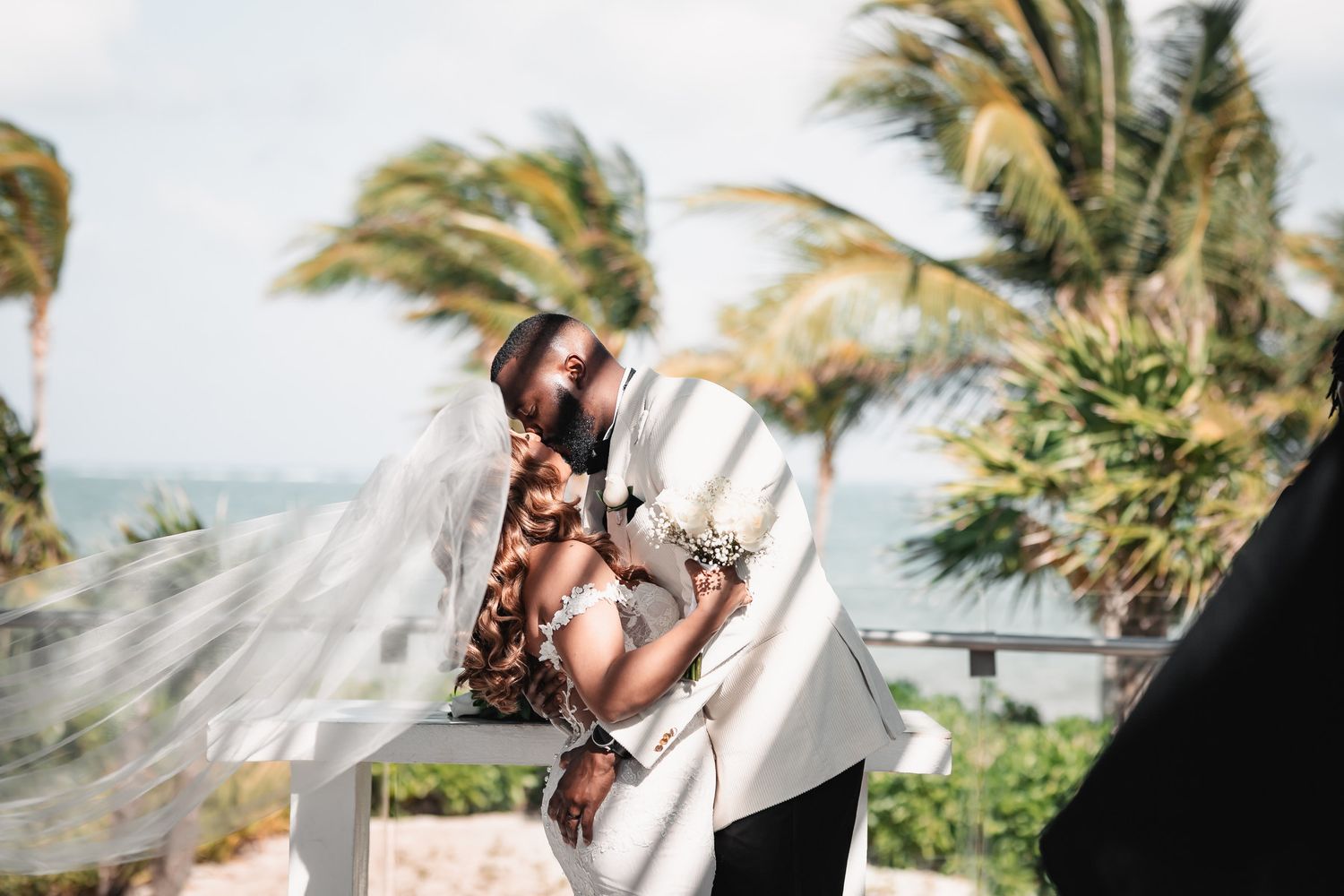 Bride and groom sharing a romantic kiss outdoors with palm trees and ocean backdrop.