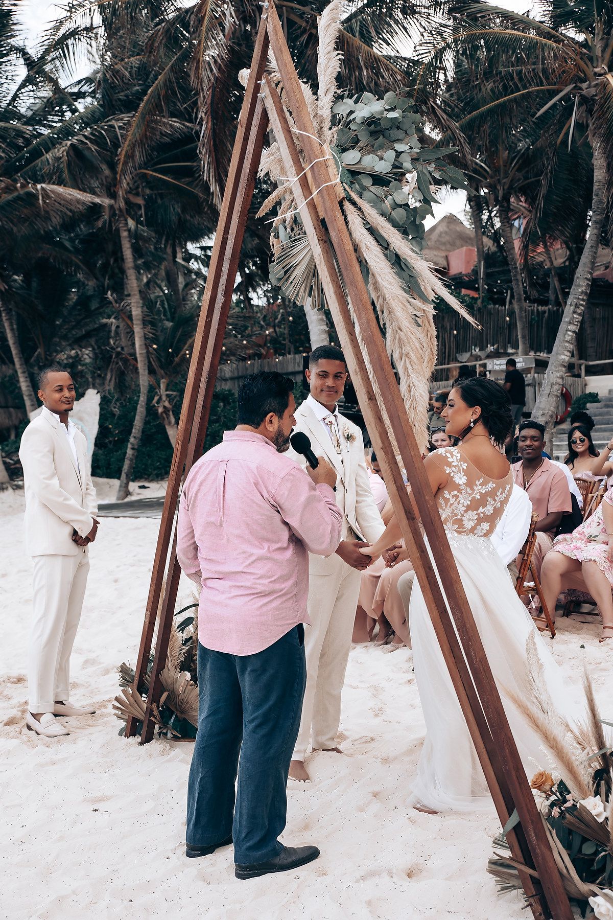 Wedding party gathers on a tropical beach with wooden structures and palm trees in the background.