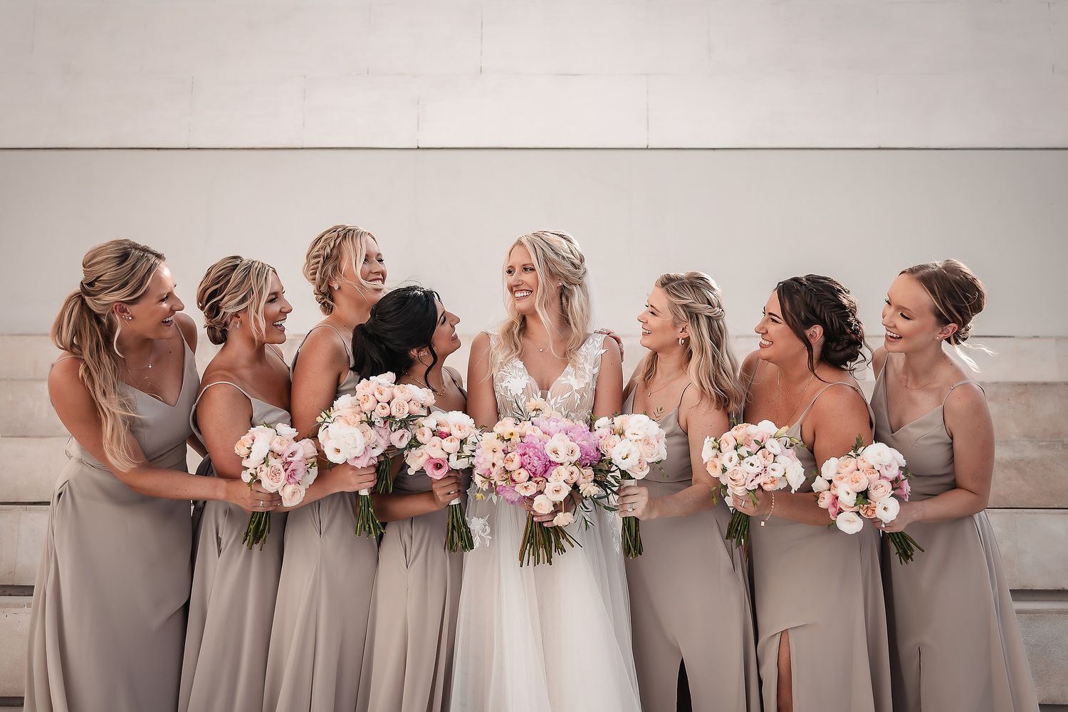 Row of bridesmaids in neutral dresses holding pink and white bouquets against light backdrop.