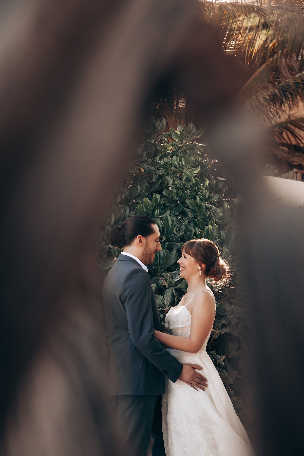 A romantic wedding couple embraces in a garden setting framed by blurred foliage in the foreground.