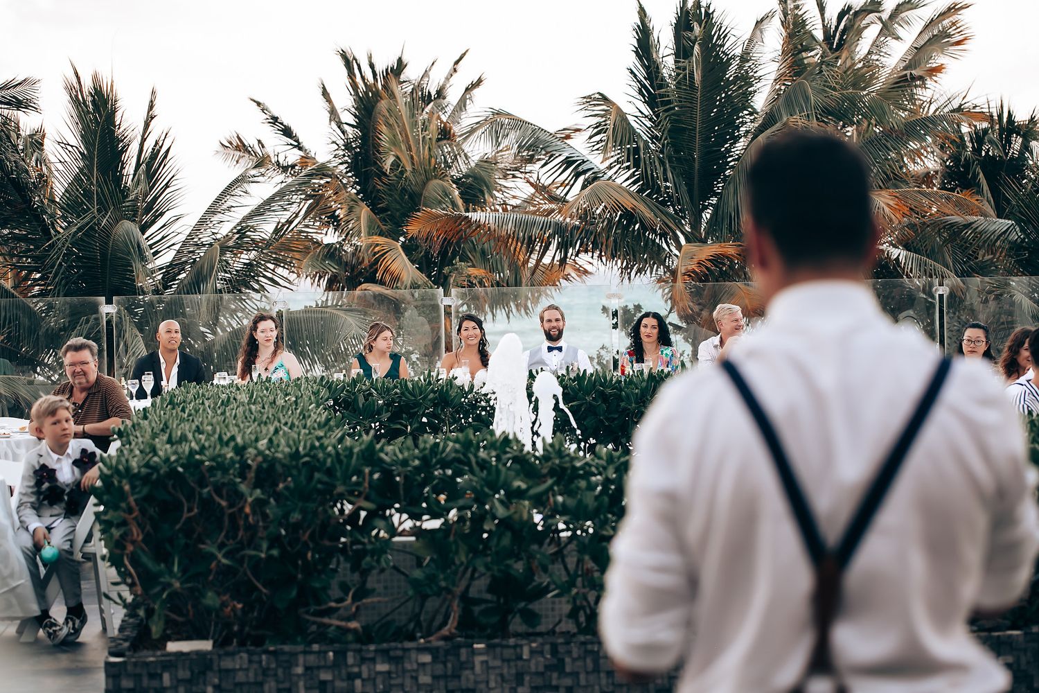 Wedding guests gather outdoors under swaying palm trees at a beachside celebration.