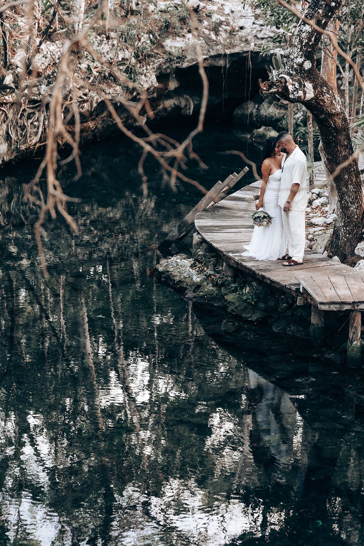 Couple poses for romantic photos by a reflective pond surrounded by trees.