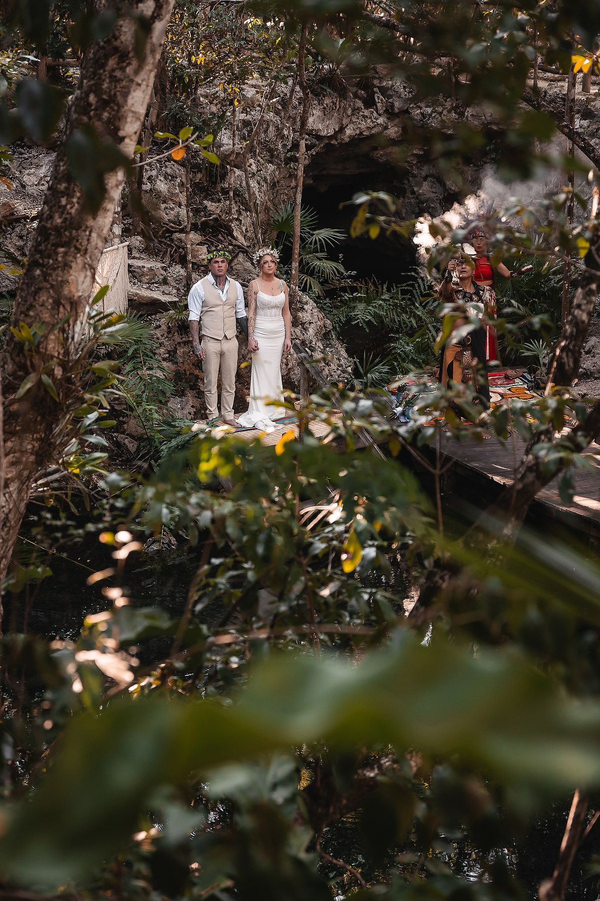 Bride and groom stand among lush tropical greenery near a cave entrance during an outdoor jungle wedding ceremony.