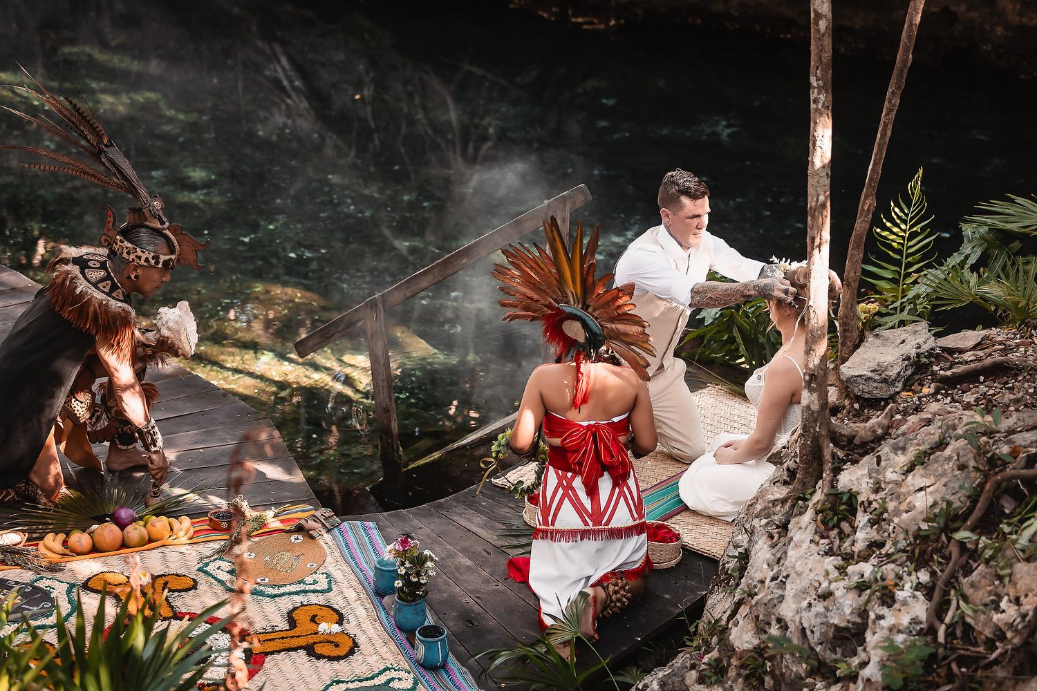 Mayan ceremony in a cenote with officiant, couple, and shaman in traditional feathered headdress amid ritual offerings.
