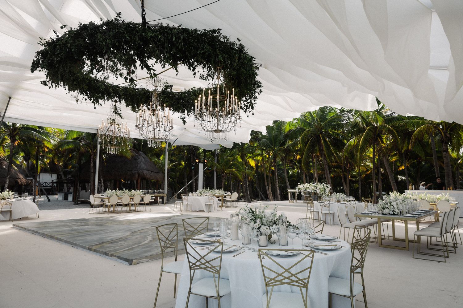 Elegant outdoor wedding reception under white tent with circular greenery chandelier and tropical palm trees.