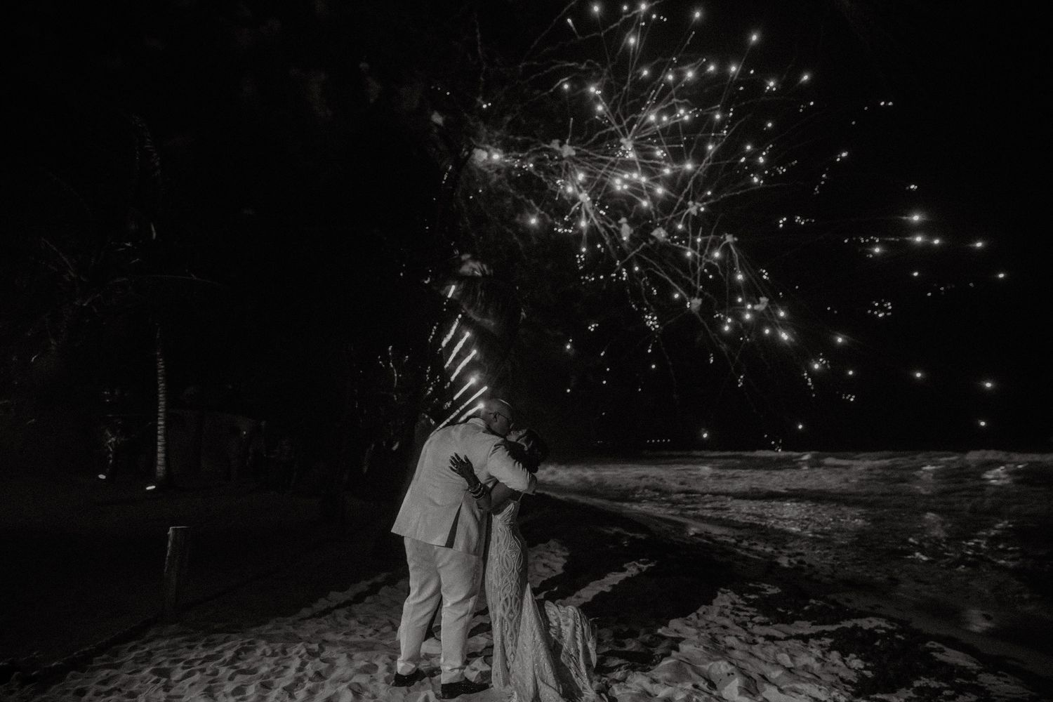 Romantic black and white photo of newlyweds embracing under fireworks on beach.