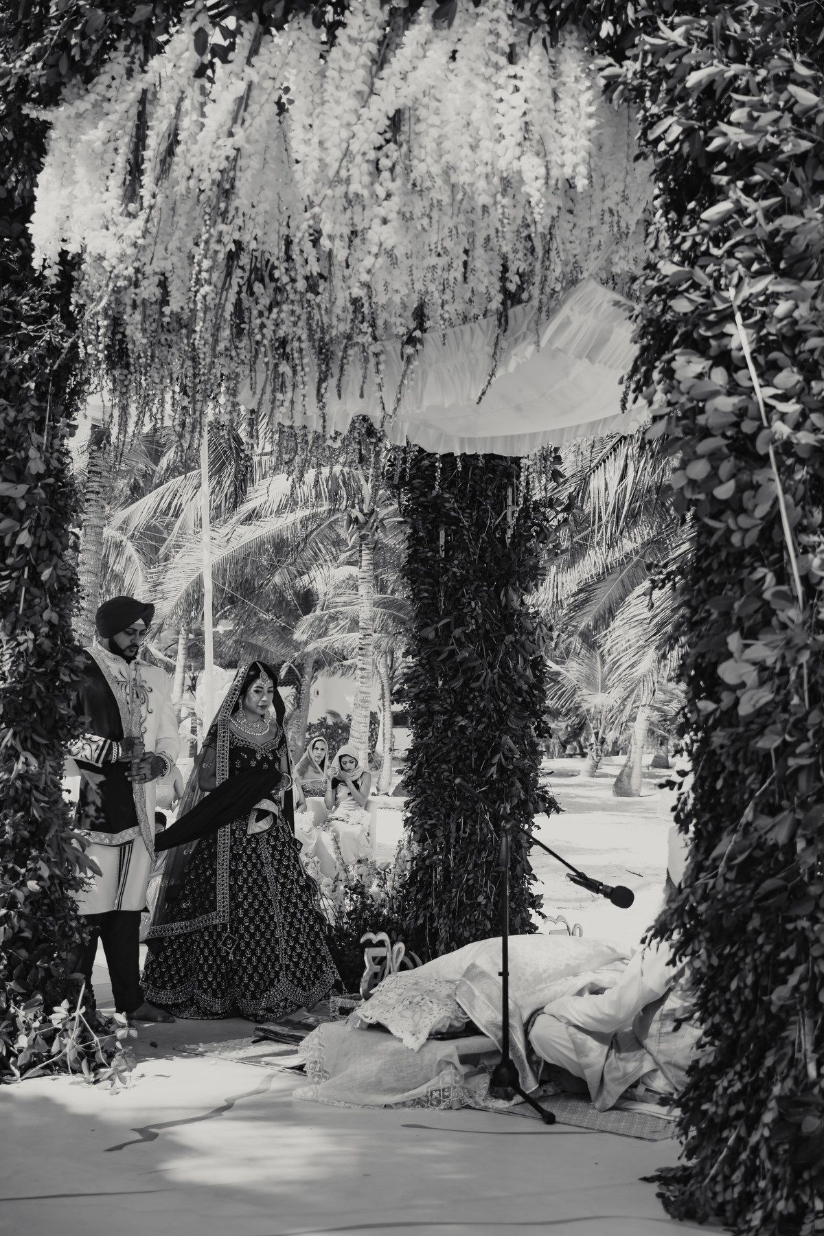 Black and white photo of bride and groom standing under floral arch during outdoor Sikh wedding ceremony.
